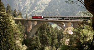 Re 460 steigt mit ihrem IR-Zug Richtung Gotthard durch den Viadukt. Fotografiert durch ein 'Fenster' des Strassentunnels