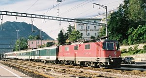 Schnellzug fährt Richtung Gotthard ab, bespannt mit einer Re 4/4 II-er. Im Hintergrund der Monte San Salvatore. 